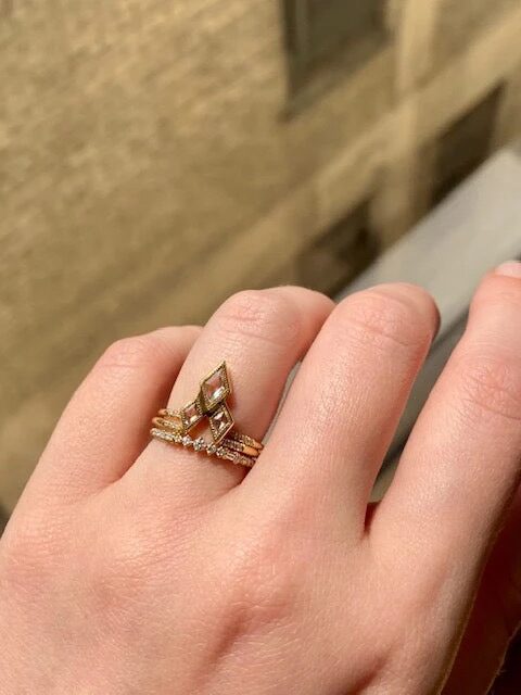 A hand with a stack of rings, including a large marquise-shaped one, in natural light near a window with a brick wall backdrop.