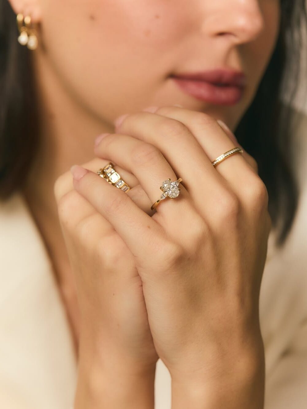 Person with short dark hair displaying hands adorned with multiple gold rings, one featuring a large diamond. Wearing small gold hoop earrings and a light-colored top.