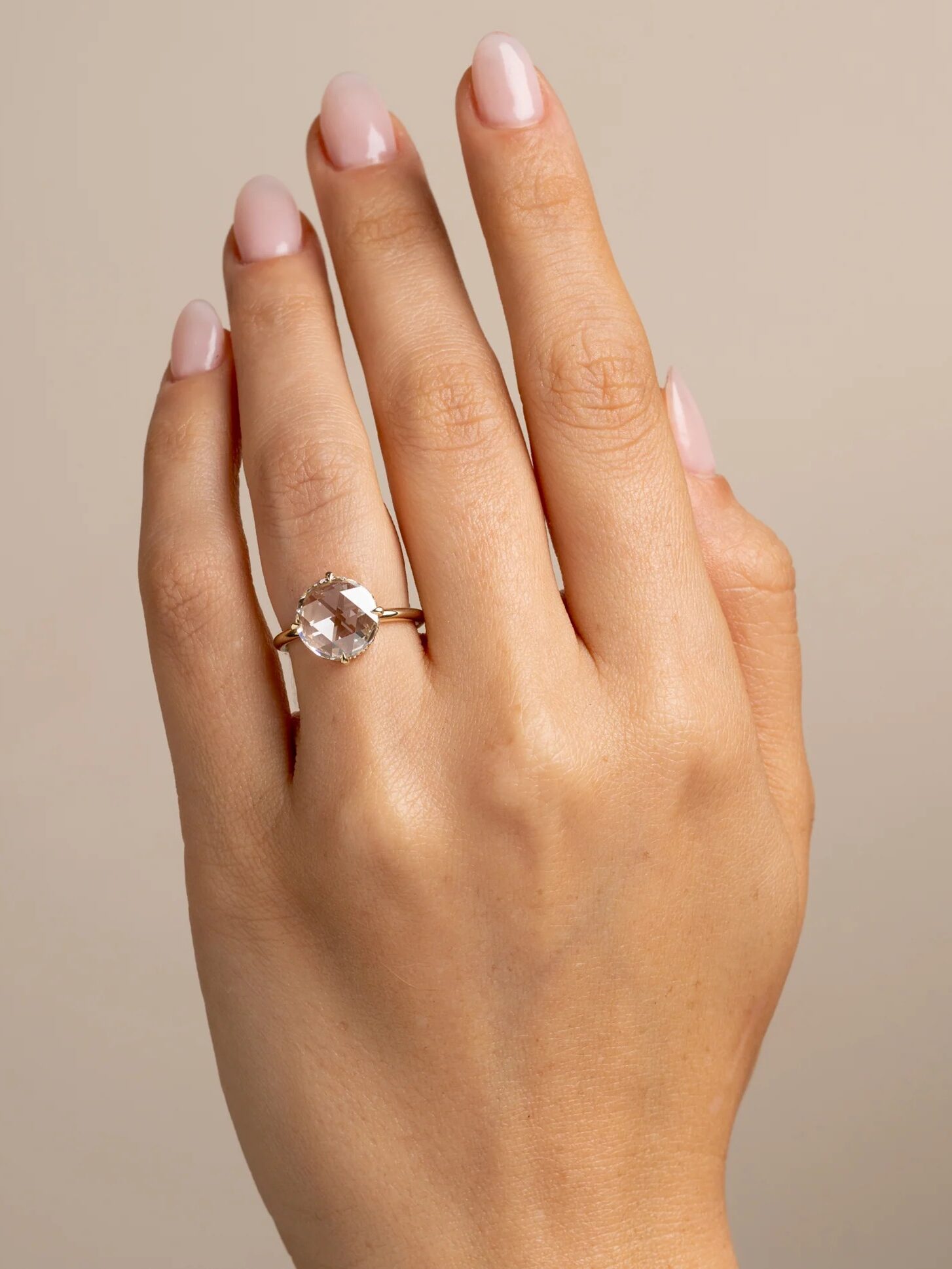 Hand with manicured nails wearing a gold ring with a large round gemstone on the ring finger, against a neutral background.