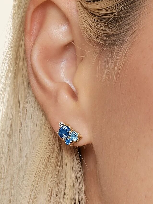 Close-up of a person’s ear wearing a gold earring set with blue and clear gemstones. The person has light skin and blonde hair.