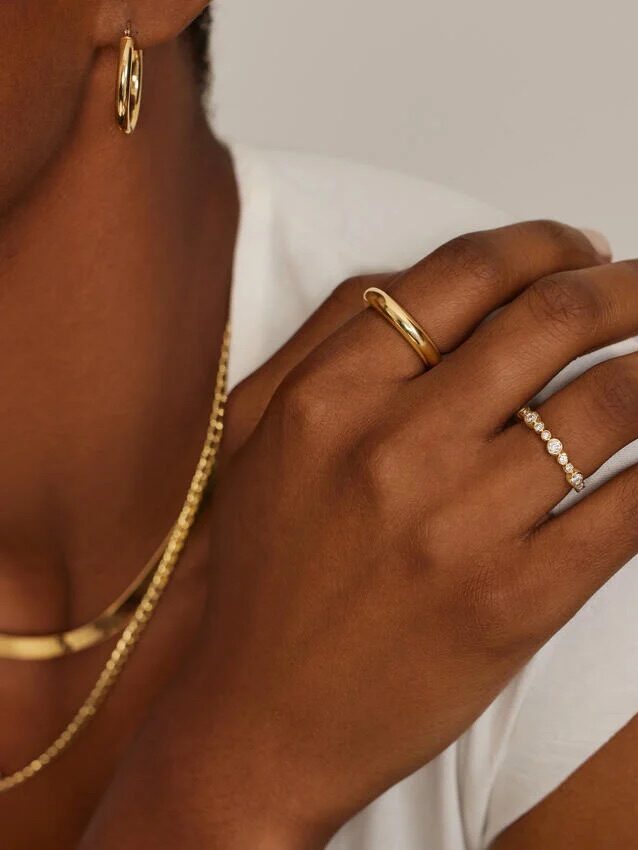 A person wearing a white shirt displays gold hoop earrings, a gold chain necklace, a gold band ring, and a diamond-studded ring on their hand.