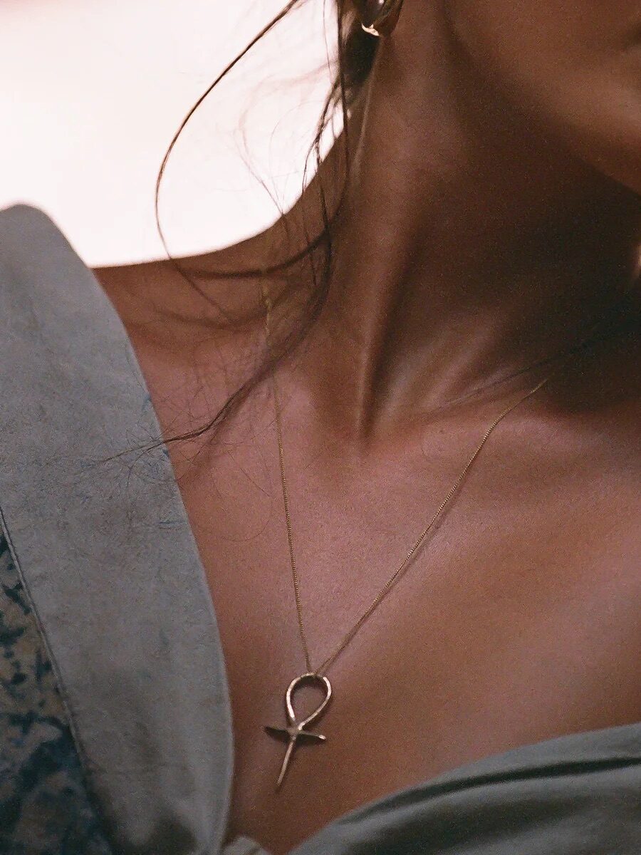 A person wearing a hoop earring and an ankh pendant necklace, with an off-the-shoulder top exposing the collarbone and shoulders.