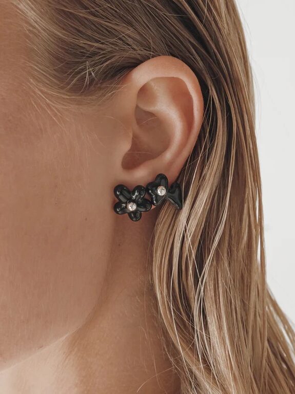 Close-up of a person’s ear wearing a black flower-shaped earring with a small pearl in the center, with wet, slicked-back hair and a neutral background.