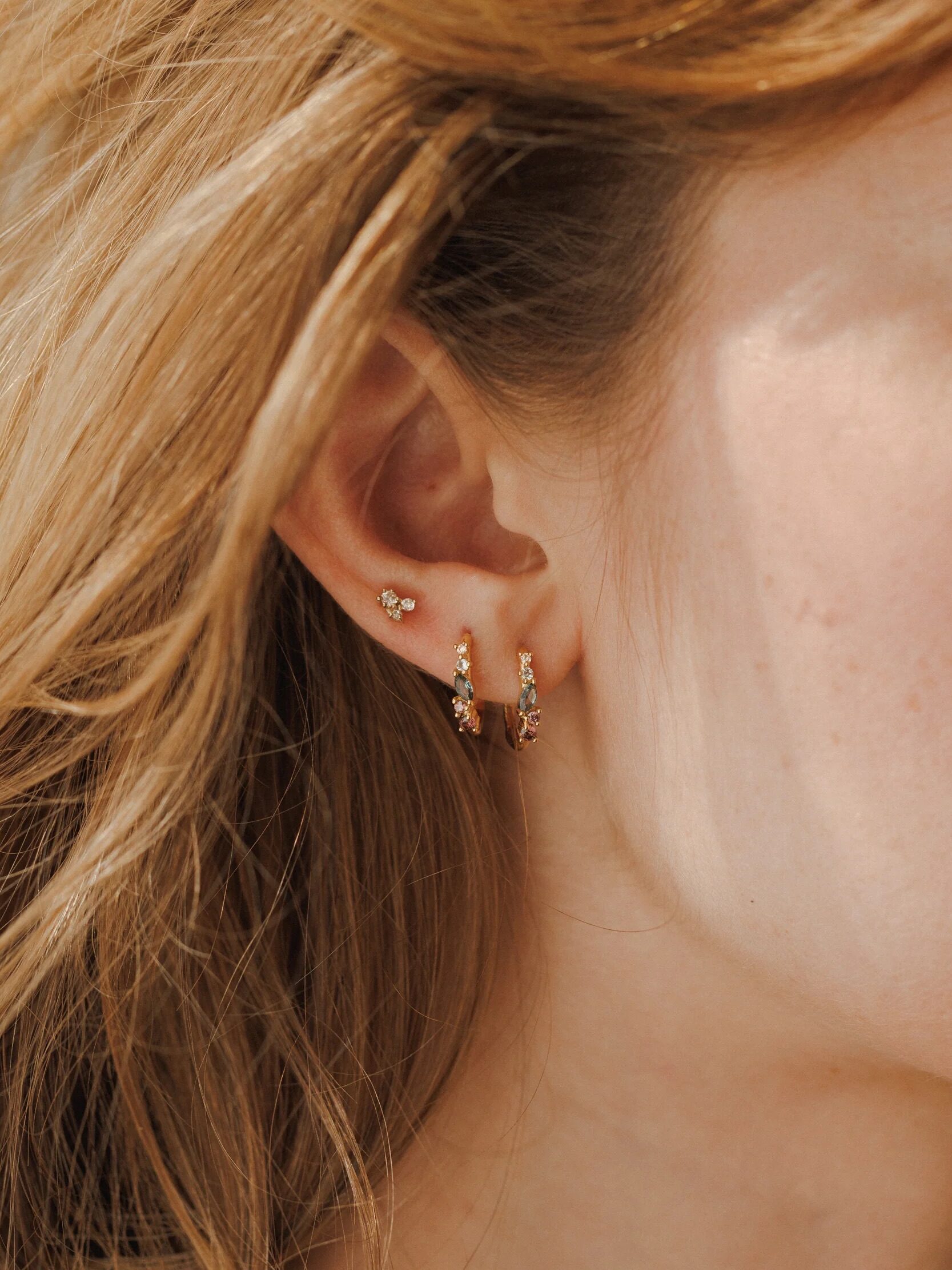 Close-up of a woman's ear with three earrings: a small stud, a medium hoop with gemstones, and a larger hoop with multiple stones, with light brown hair partially visible.