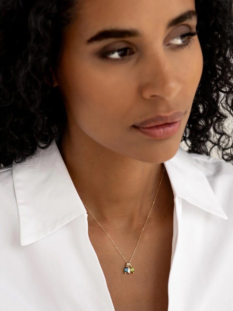 A woman with curly hair wearing a white collared shirt and a delicate necklace with a small pendant looks to the side.