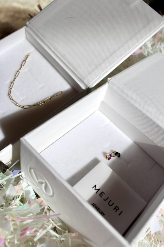 Two open white jewelry boxes; one contains a gold chain necklace, and the other holds a small ring with a purple stone and a Mejuri jewelry card.