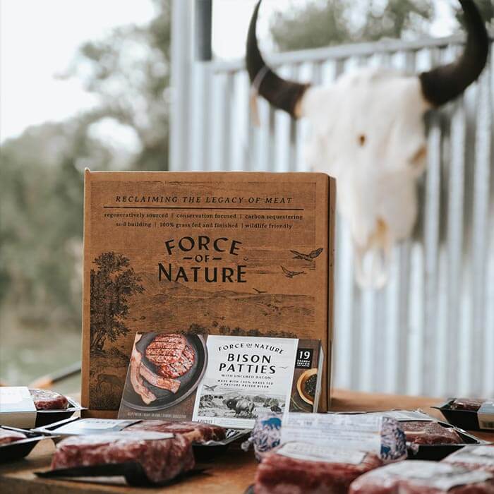 A box labeled "Force of Nature" and a package of bison patties are surrounded by various packed meat products. A cow skull is mounted on a wall in the background.