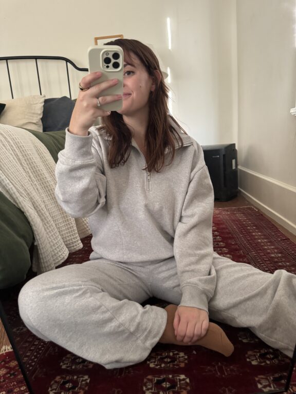 A person in a gray sweatsuit sits cross-legged on a red patterned rug, taking a mirror selfie in a bedroom with a bed and blanket visible in the background.