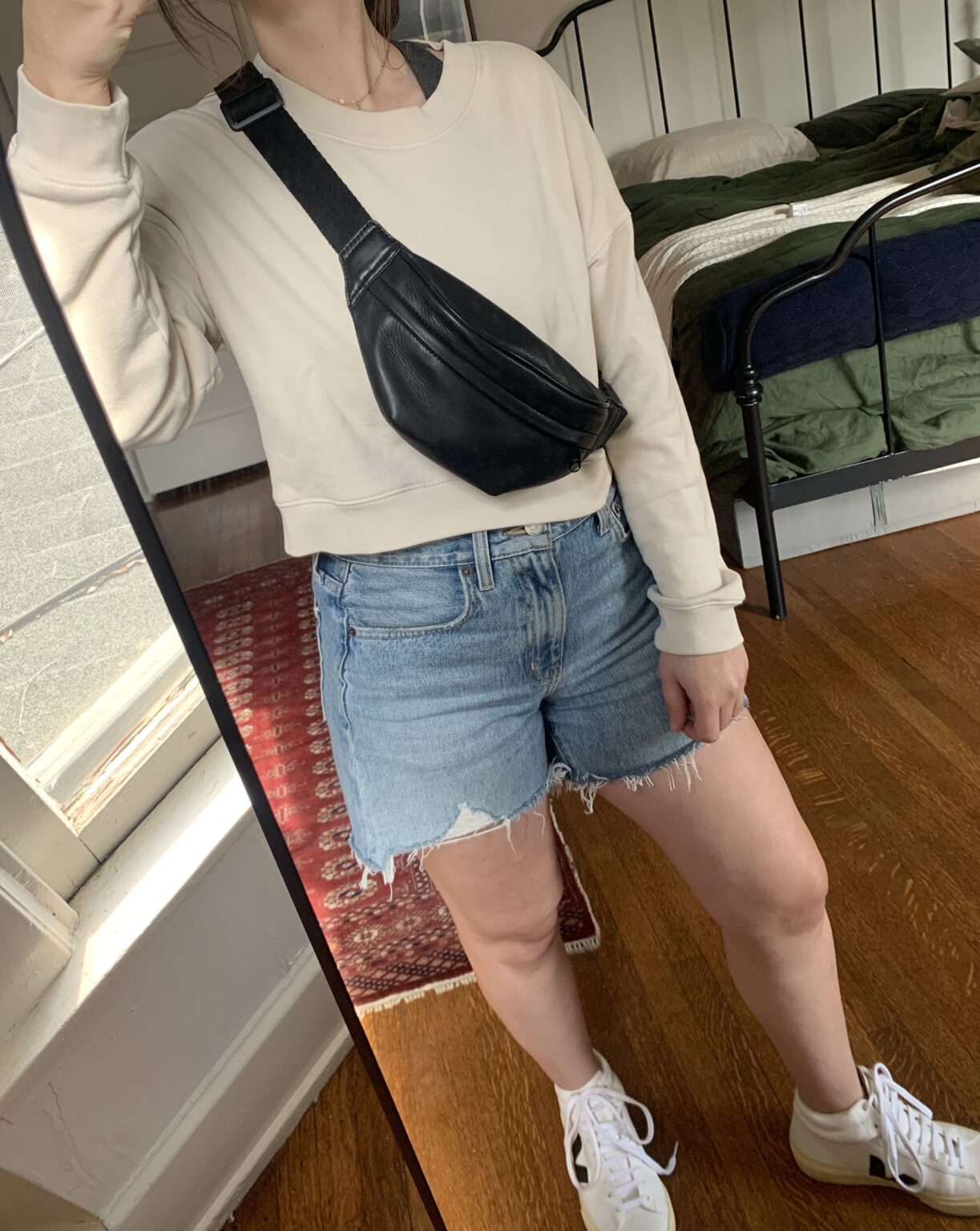 Person taking a mirror selfie wearing a cream sweatshirt, denim shorts, white sneakers, and a black crossbody bag in a bedroom with wooden floors and a made bed.