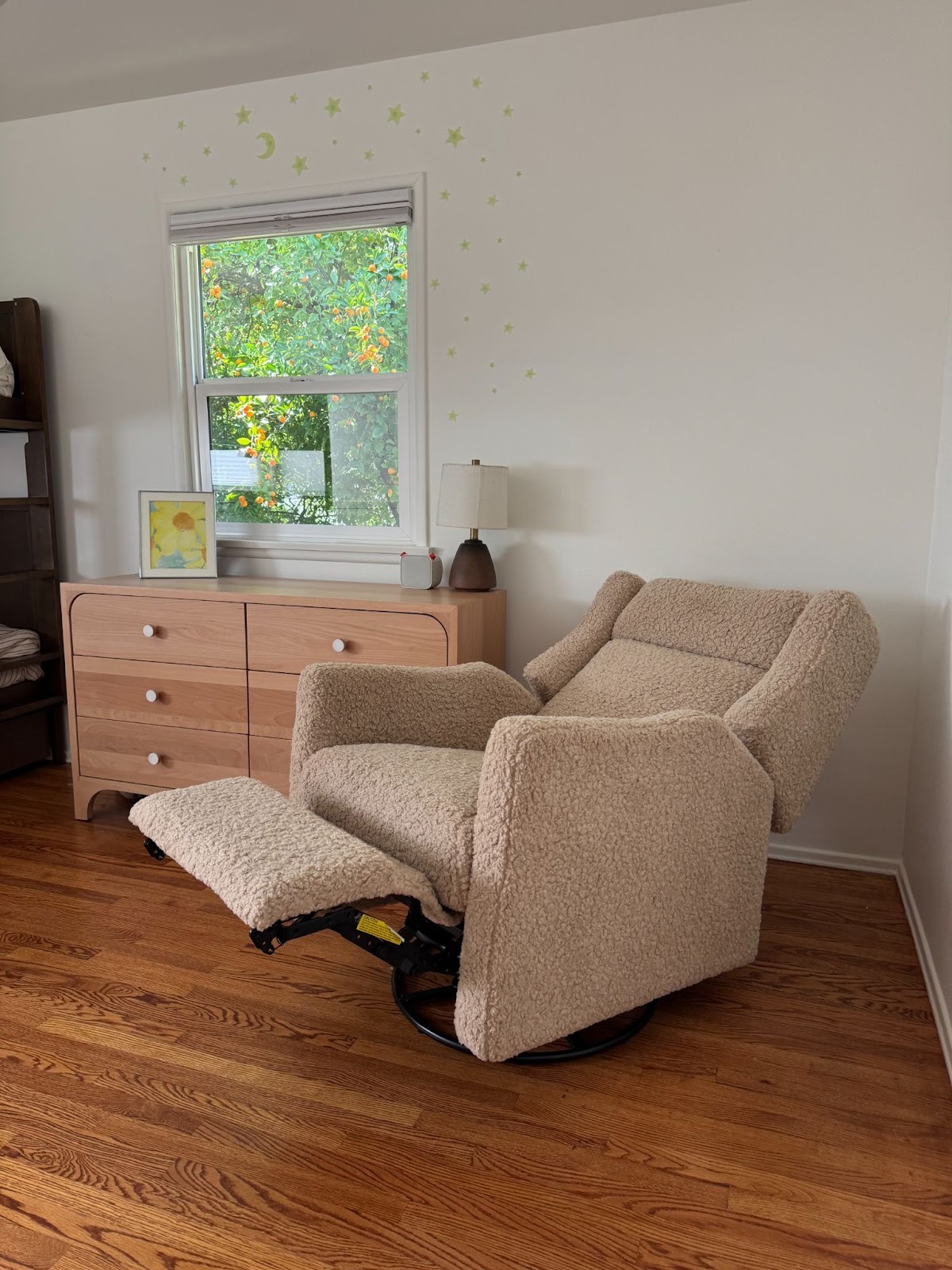 A beige recliner chair with an extended footrest is placed in a room with wooden floors, next to a light wood dresser and a window with green foliage outside.