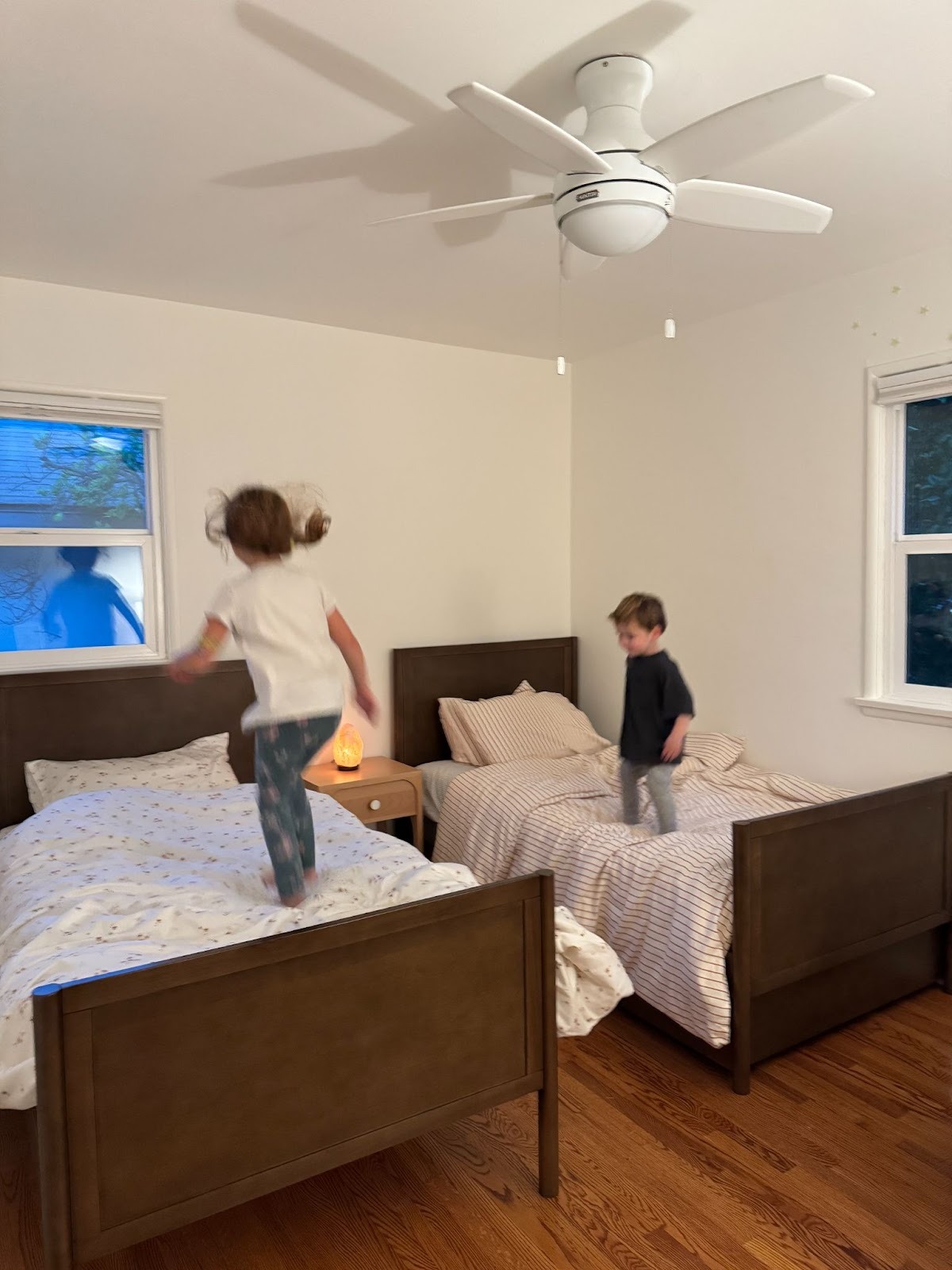 Two children are playing in a bedroom with wooden beds, one jumping on a bed and the other standing beside a bed. There is a ceiling fan and windows with natural light.