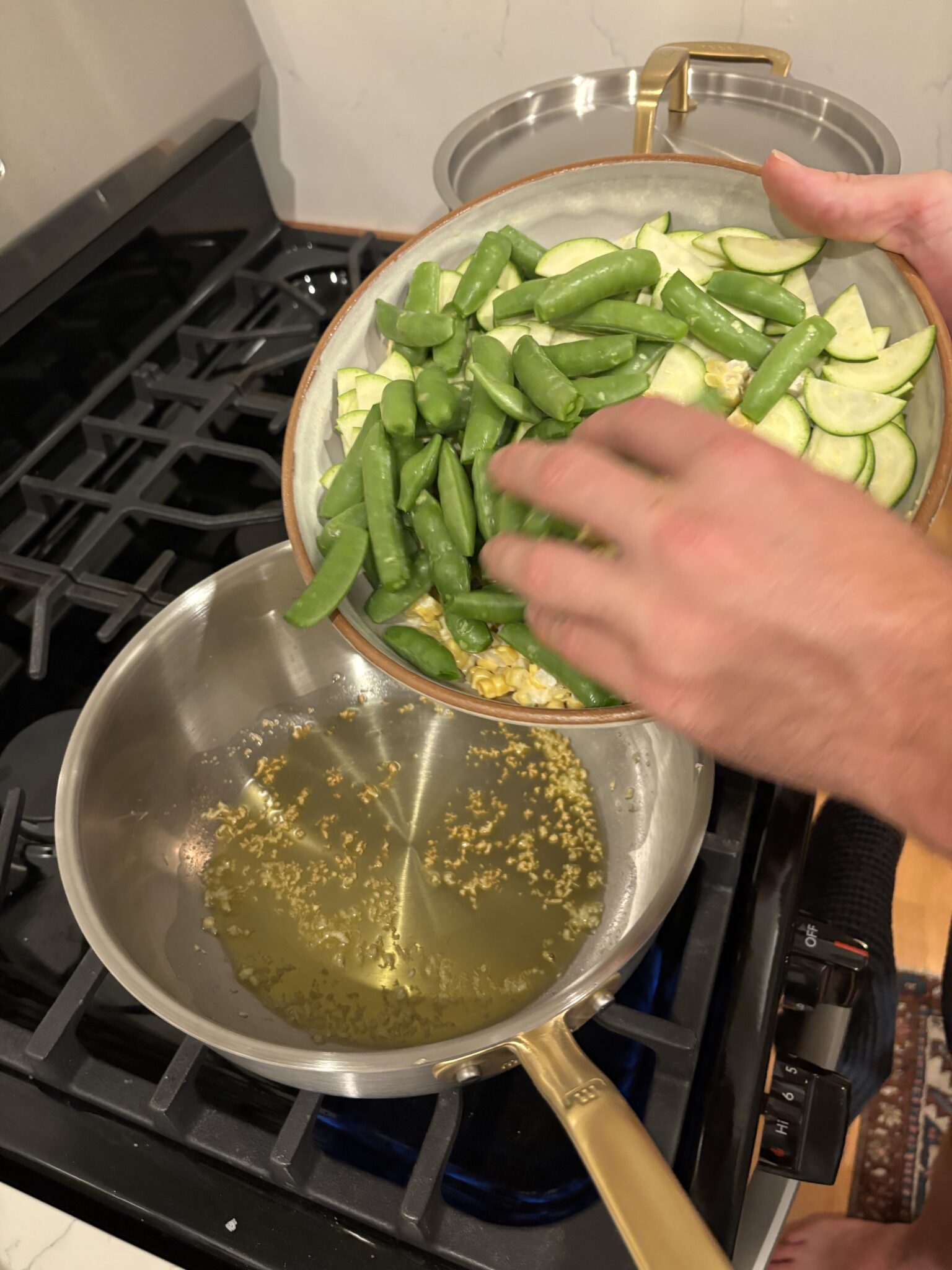 A person adds sugar snap peas and sliced zucchini from a bowl into a saucepan with sizzling garlic and oil on a stove.