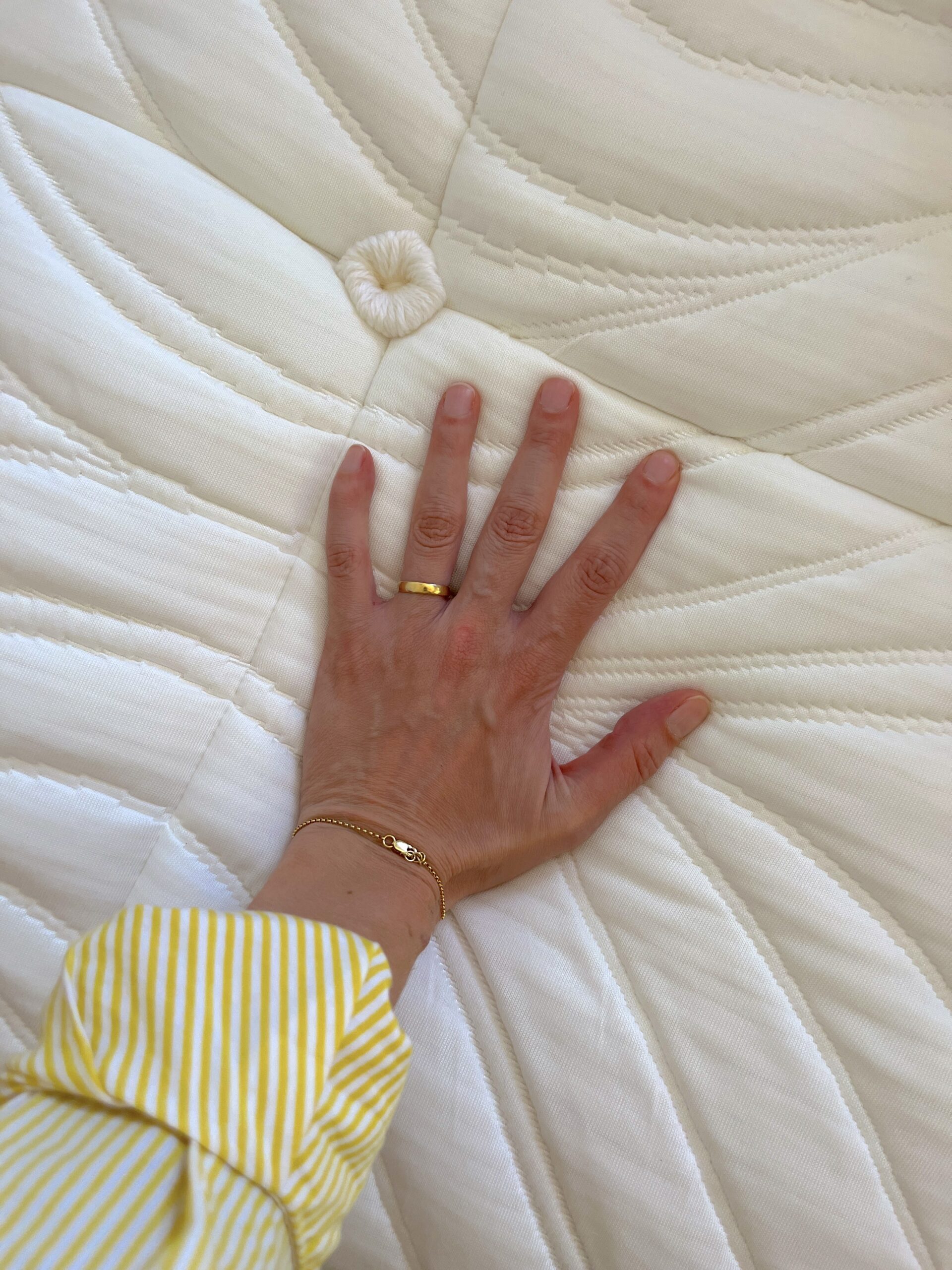 Ashley D'Arcy's hand has a gold ring and bracelet, and rests on the quilted fabric surface of a Brentwood Home organic mattress, with a yellow striped sleeve visible.