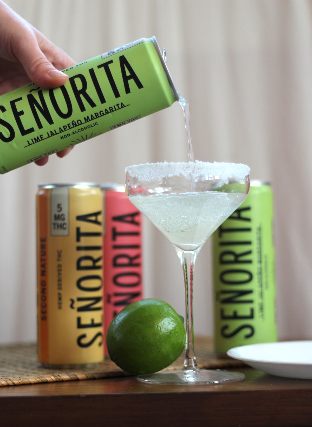 A can of Señorita lime jalapeño margarita is being poured into a sugar-rimmed glass, with other cans and a lime nearby.