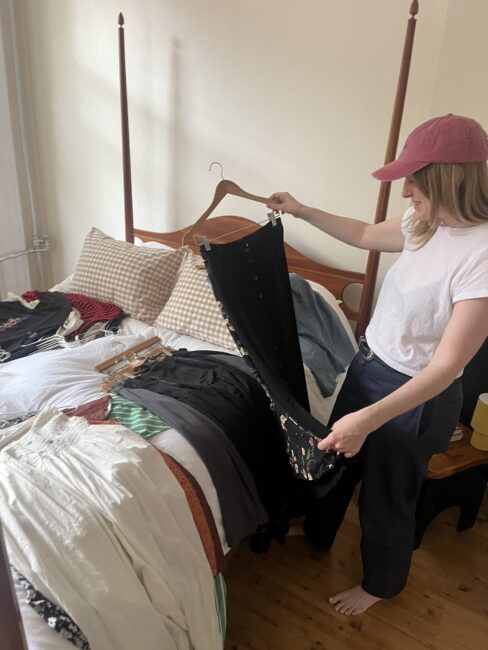 A person wearing a pink cap and white t-shirt stands by a bed holding up a black garment, with clothes spread out on the bed.