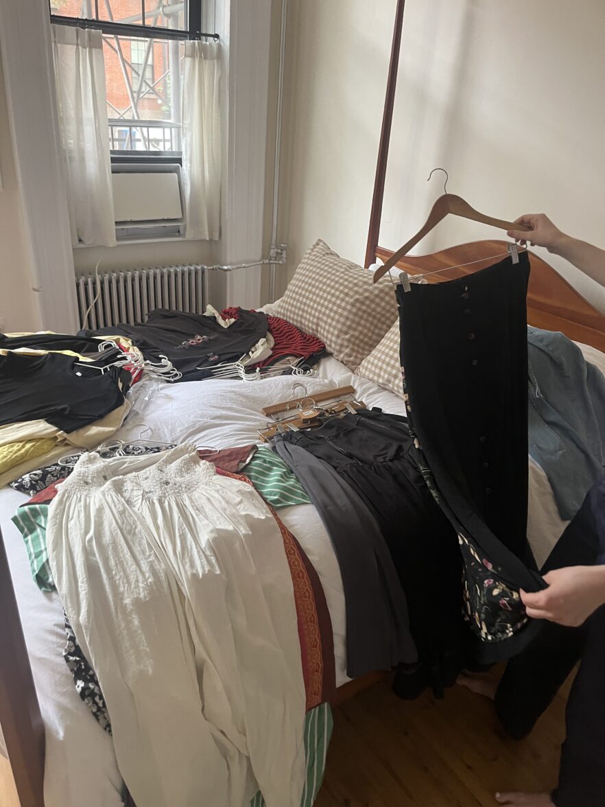 A person holds up a pair of black pants over a bed covered with folded and spread out clothing in a sunlit bedroom.