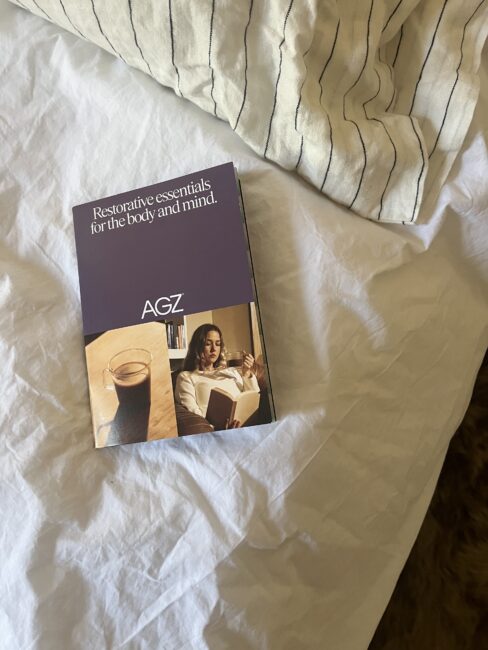 A brochure titled "Restorative essentials for the body and mind" with a photo of a woman reading and a cup of coffee rests on a white, unmade bed with a striped pillow.