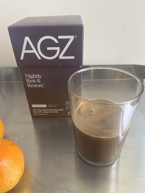 A glass of chocolate beverage sits next to an AGZ Nightly Rest & Restore chocolate supplement box on a metal surface, with two oranges nearby.