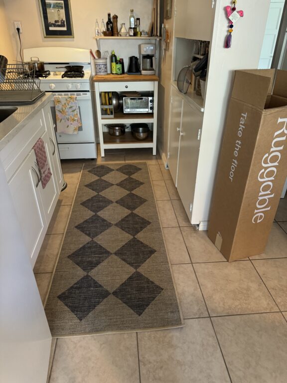 A narrow kitchen with tiled floor, patterned runner rug, stove, coffee maker, microwave, and an empty Ruggable box standing upright near a white cabinet.