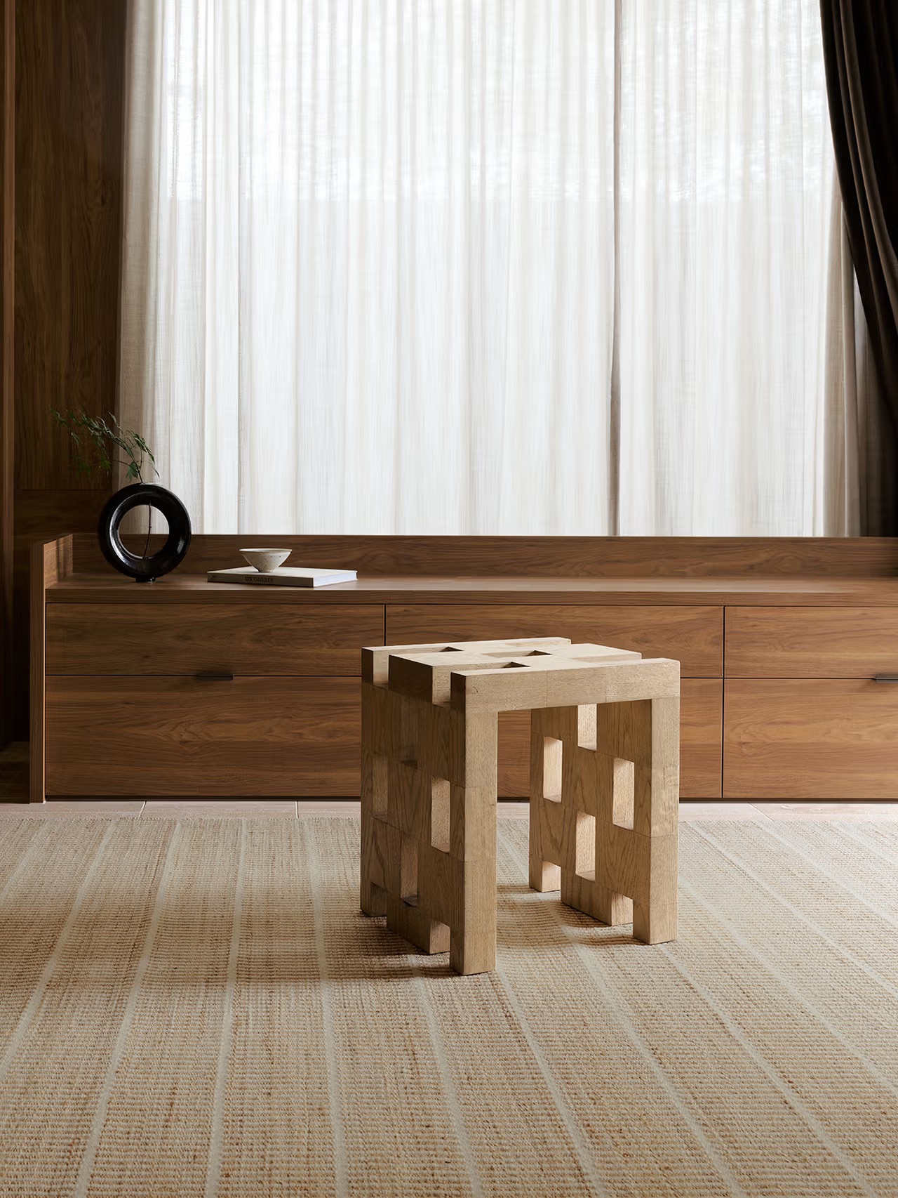 A modern wooden stool with square cutouts stands on a beige rug in front of a low wooden cabinet and sheer curtains in a minimalist room.