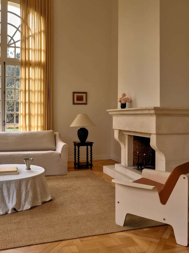 A minimalist living room with a cream sofa, round coffee table, fireplace, black side table with a lamp, and a large window with sheer curtains.