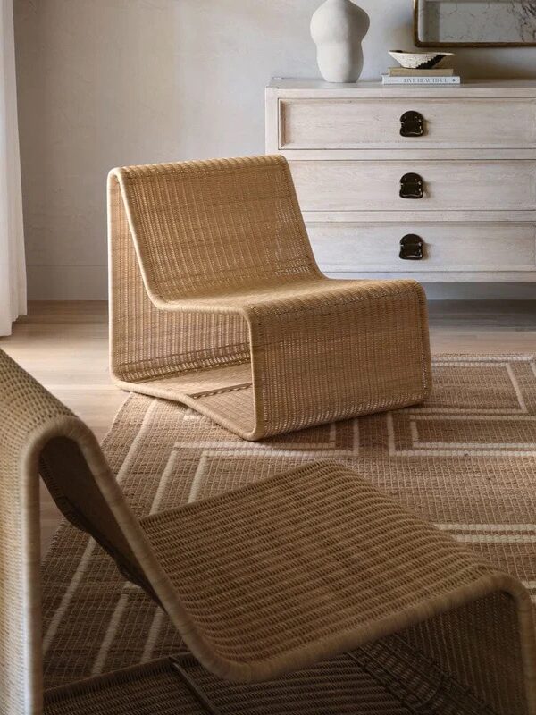Two woven rattan lounge chairs sit on a patterned rug in front of a light wood dresser with black handles, in a minimal, modern room.