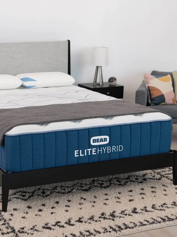 A modern bedroom featuring a Bear Elite Hybrid mattress on a black bed frame, two pillows, a gray throw blanket, and an armchair; a bedding award badge is displayed in the foreground.