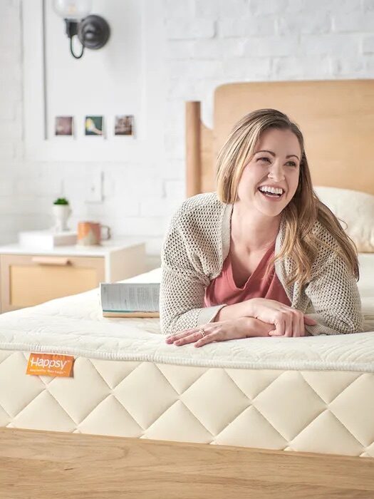 A woman lies on a bed, smiling and holding an open book. The mattress has a quilted pattern and a visible Happsy label. The room has a white brick wall and wooden furniture.
