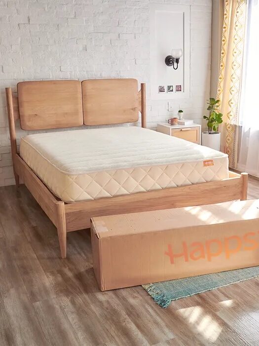 A minimalist bedroom with a wooden bed frame, mattress, bedside table, and a large box labeled "HAPPSY" positioned at the foot of the bed.