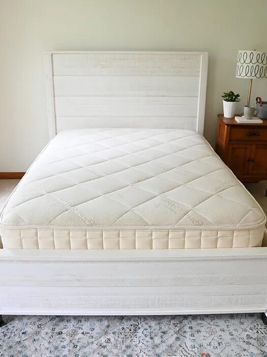 A neatly made bed with a light-colored mattress on a white wooden bed frame, next to a wooden nightstand with a lamp and potted plants in a bright bedroom.