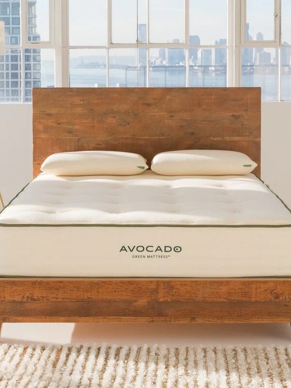 A wooden bed frame with an Avocado Green Mattress and two pillows in a bright room with large windows and a city skyline view.