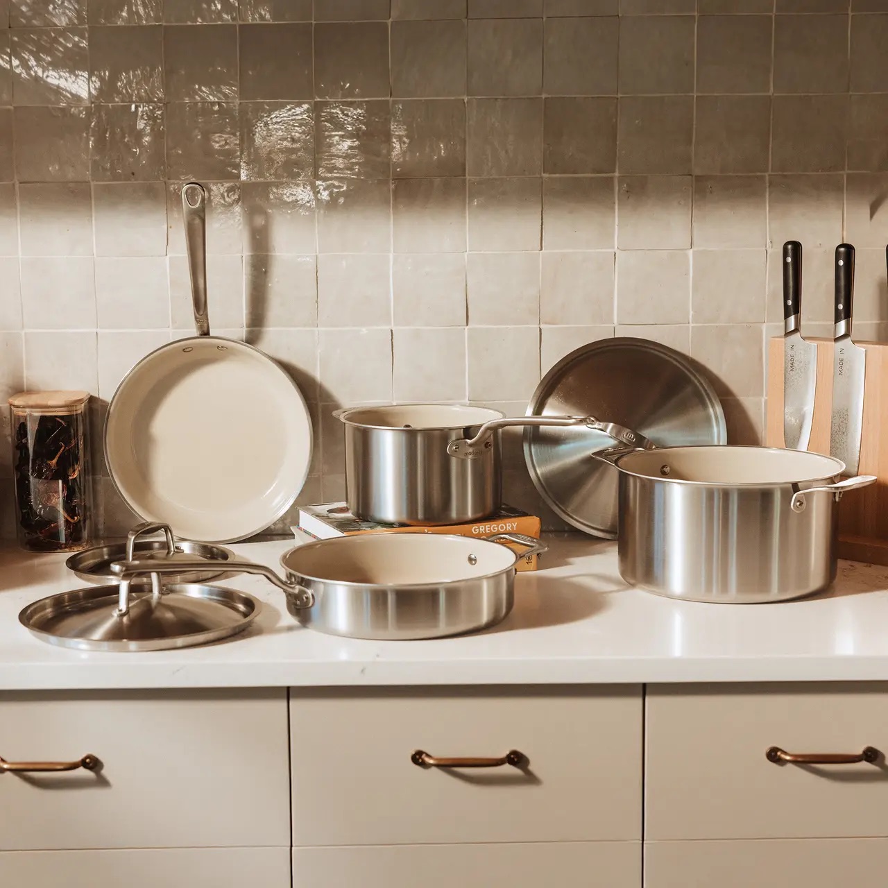 A set of stainless steel pots, pans, and lids is arranged on a white kitchen counter, with knives and a cutting board in the background.
