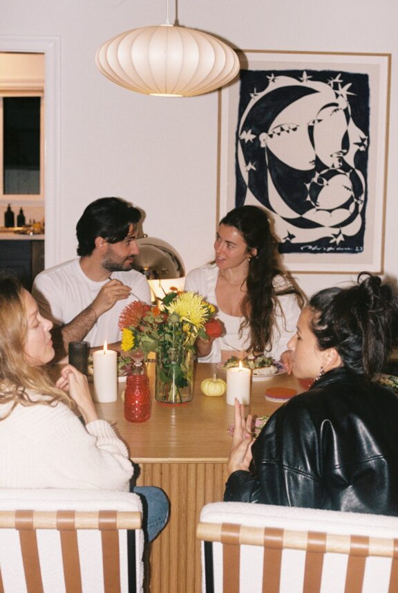 Four people sit around a wooden dining table with flowers, candles, and drinks, having a conversation in a warmly lit room with artwork on the wall.
