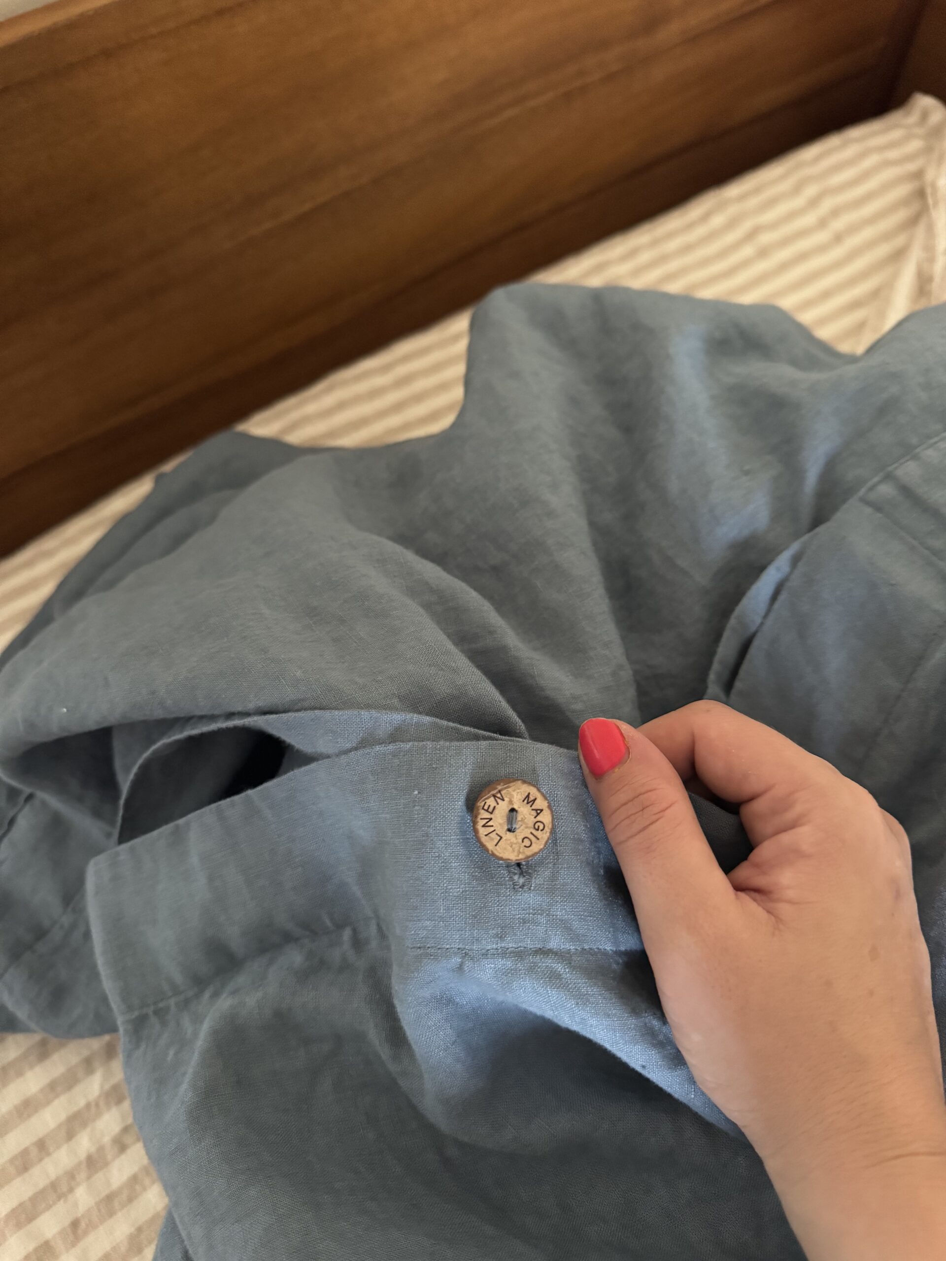 A hand with red nail polish holds a blue garment with a visible button stamped “LINEN, MADE IN FRANCE” over a striped bedsheet.