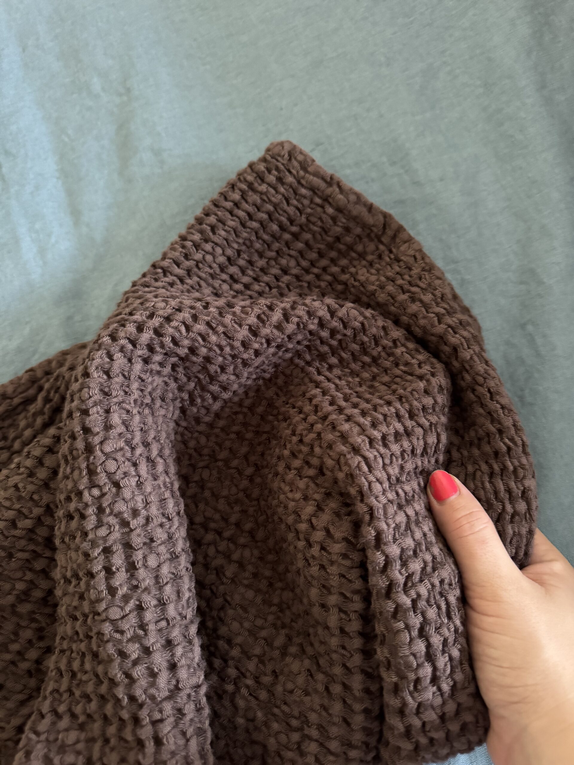 A hand with red nail polish holds a textured, brown knitted fabric against a light blue background.