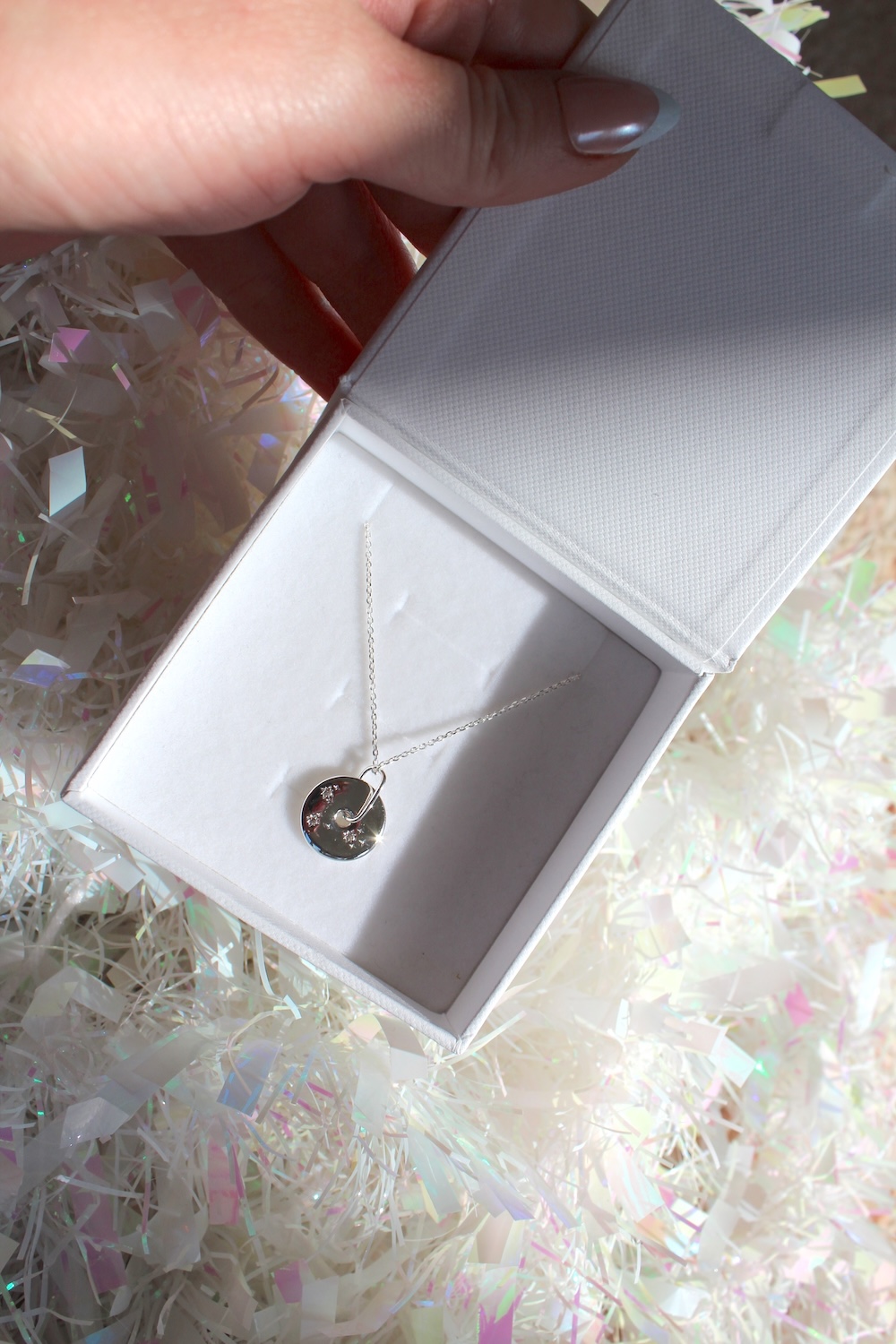 A hand holds open a white jewelry box displaying a silver necklace with a round pendant, set against a background of iridescent shredded paper.
