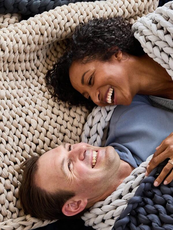 A woman and a man lie close together, smiling and wrapped in chunky knit blankets of different colors.