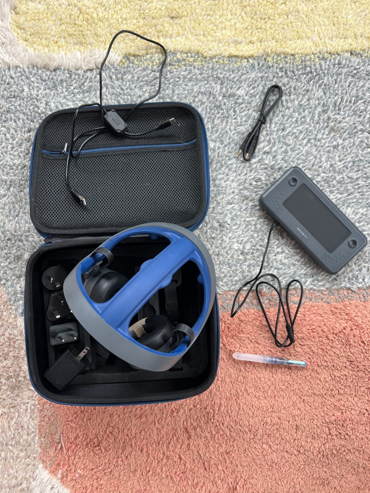 A virtual reality headset with controllers and cables is stored in an open carrying case on a multicolored rug, next to a tablet and stylus.