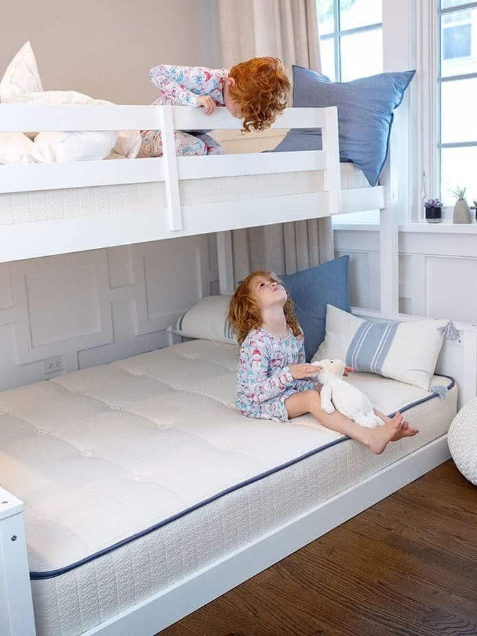 Two children in pajamas are on a bunk bed. One is on the top bunk peeking over the edge, and the other is sitting on the bottom bunk holding a stuffed toy bear, both nestled comfortably thanks to their cozy nontoxic mattresses from My Green Mattress.