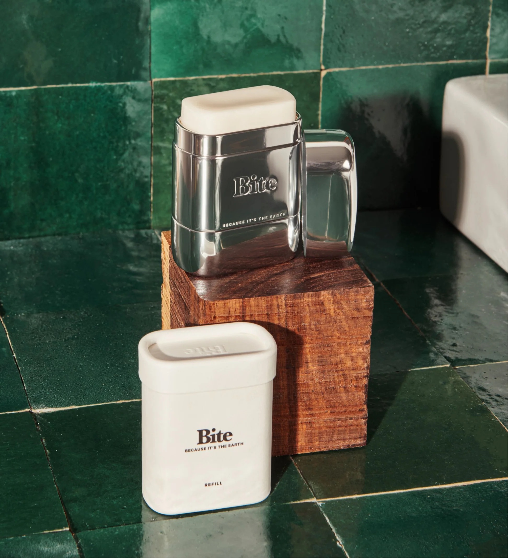 Two Bite brand containers, one stainless steel mug with a lid and one white refill container, are placed on a wooden block against a background of green tiled wall.