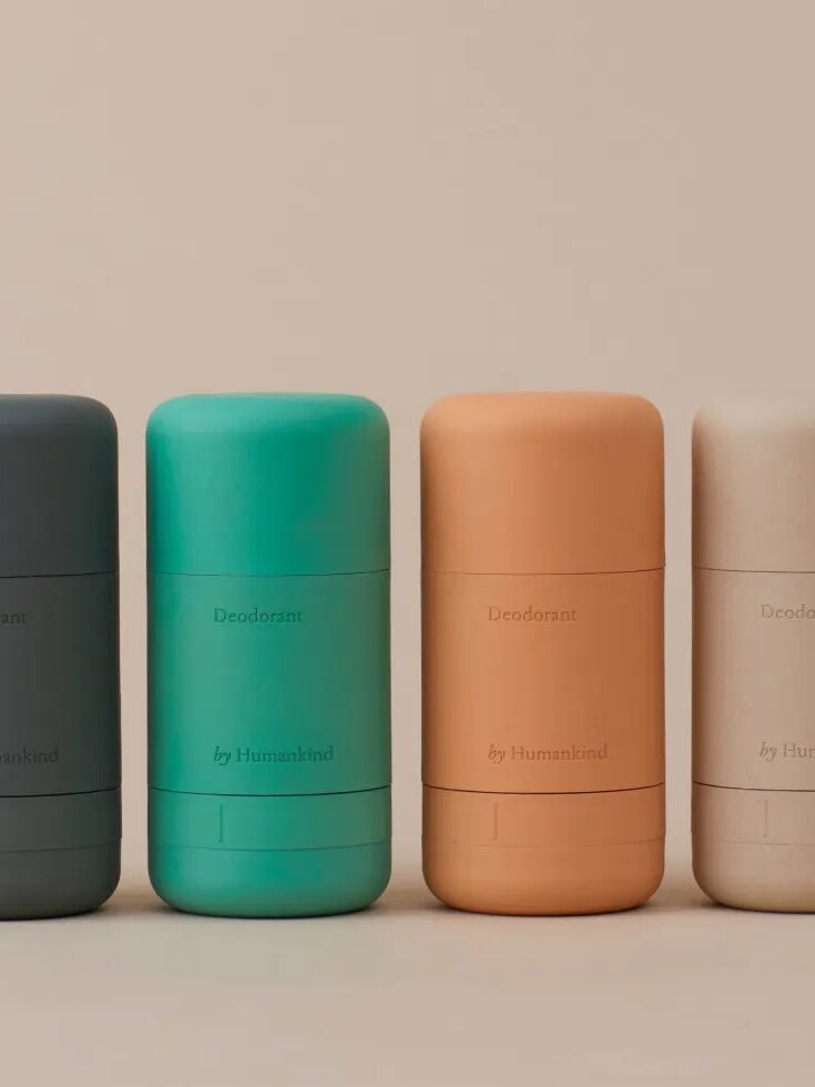 Four cylindrical deodorant sticks in grey, green, orange, and beige colors are arranged in a row on a beige background. The label on each reads "Deodorant by Humankind".