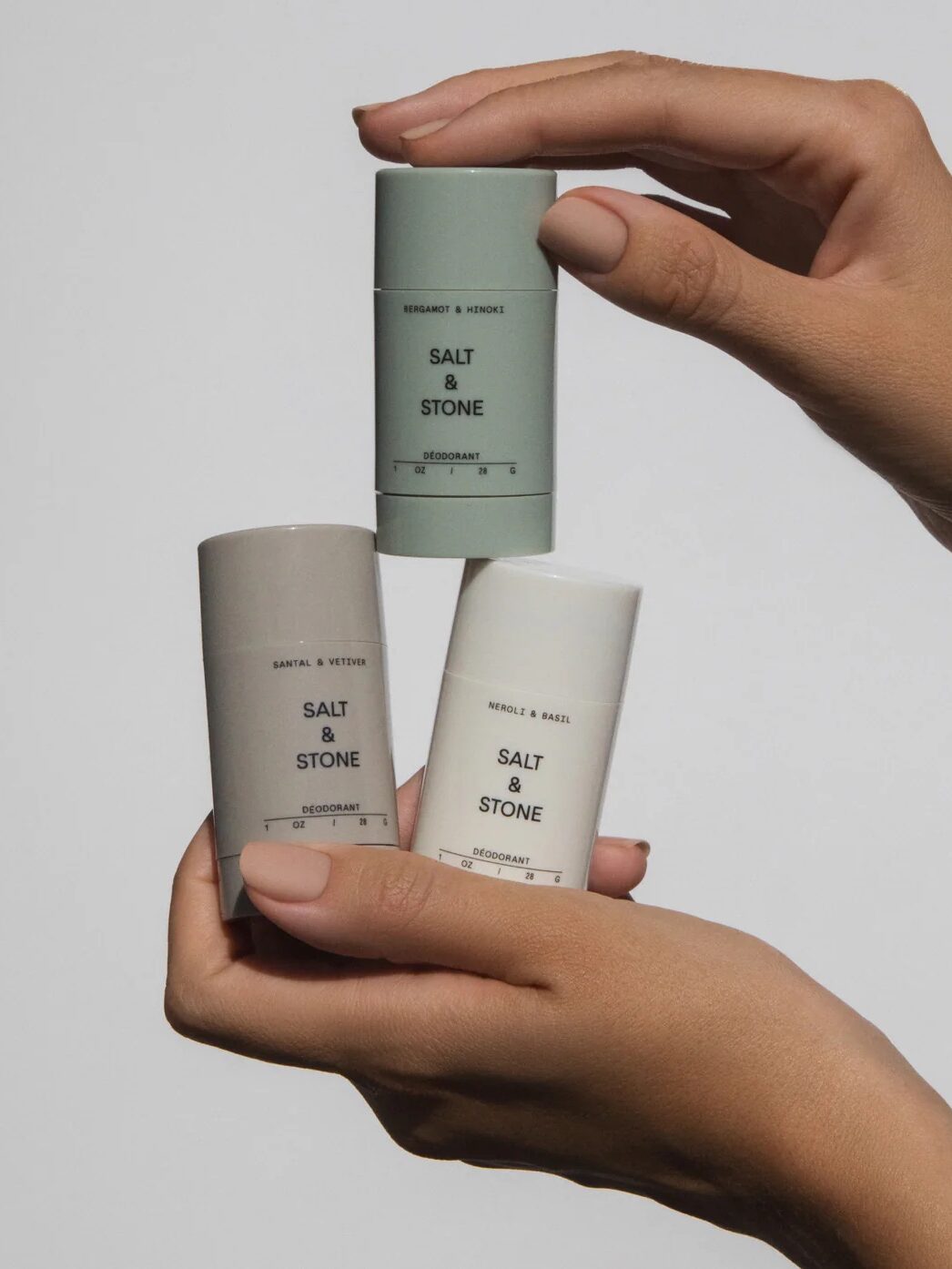 Three Salt & Stone deodorant sticks in different scents are held by hands against a plain background.