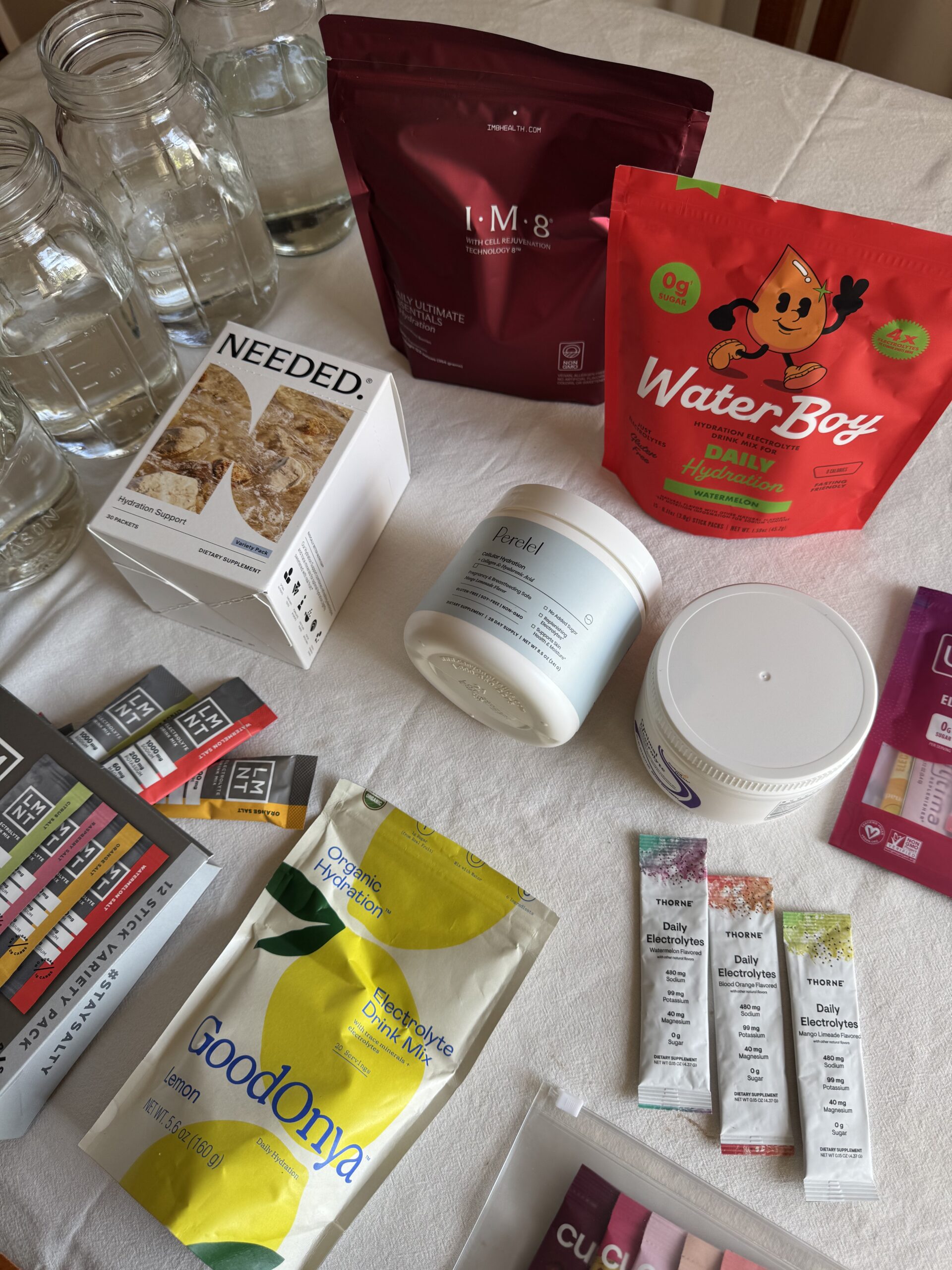 Various wellness and nutrition products, including supplements, snacks, drink mixes, and jars of water, are arranged on a white tablecloth.
