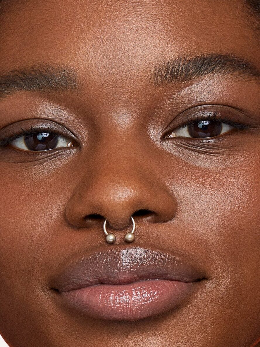 Side-by-side photos of a person with a septum piercing, showing "Before" with natural skin and "Revealed" with smoother, more even-toned makeup.