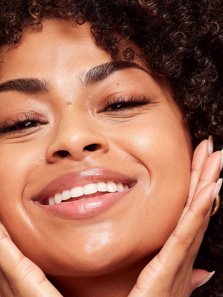 Close-up of a smiling person with curly hair, clear skin, and hands touching their face on both sides.