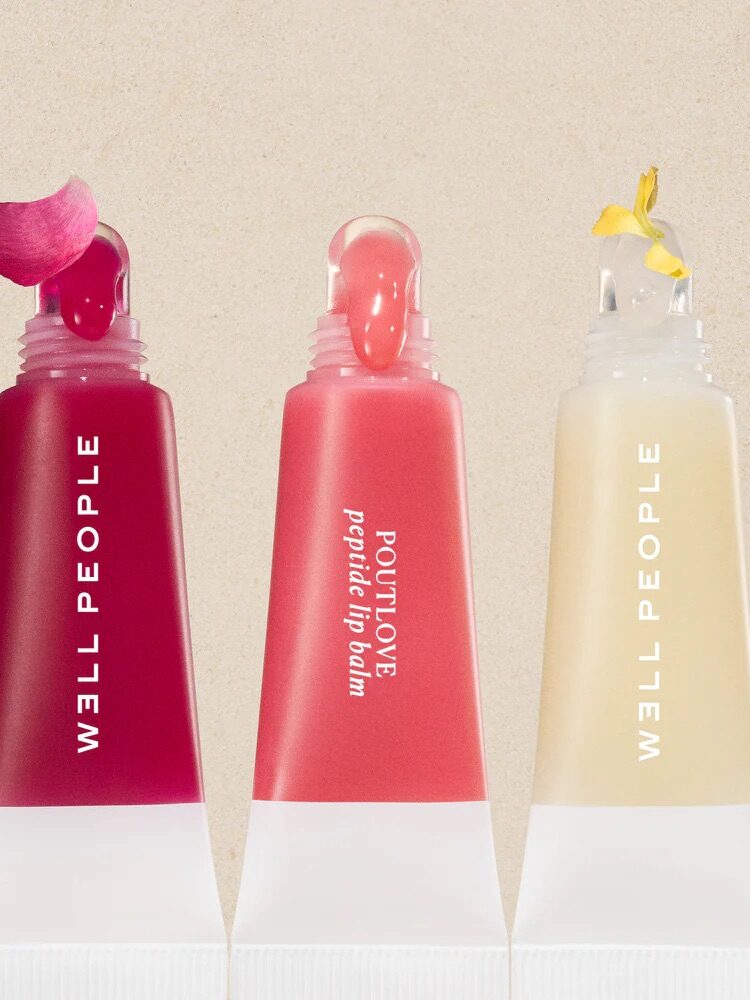 Four tubes of WELL PEOPLE Poutlove peptide lip balm in different shades, each with a small decorative flower or petal on top of the applicator.