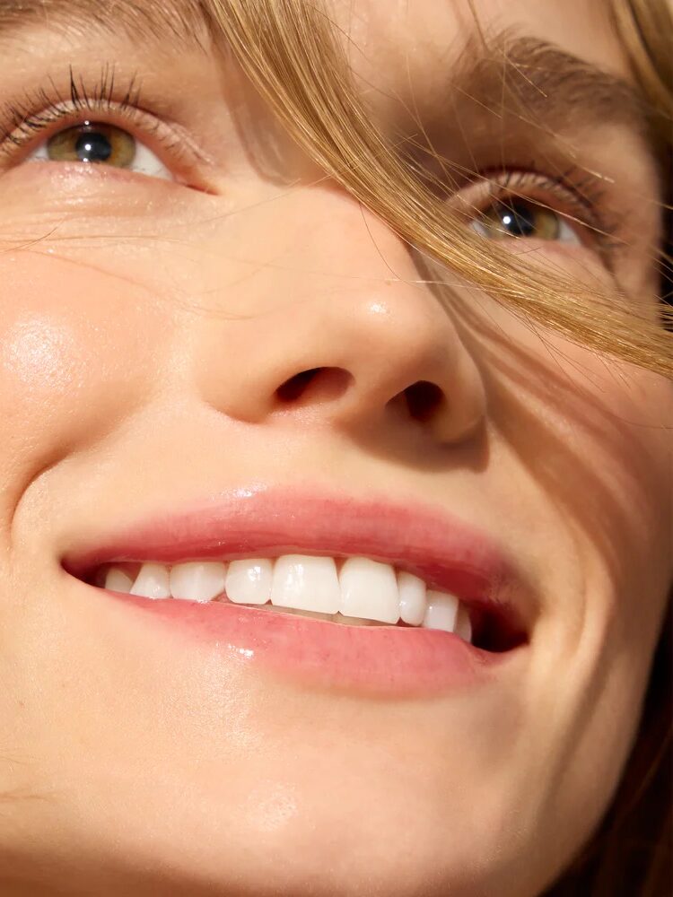 Close-up of a person smiling with clear skin, natural makeup, and a few strands of hair across their face.