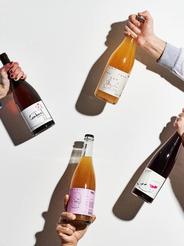 Four hands hold up different bottles of wine and cider against a plain white background, casting distinct shadows on the wall.