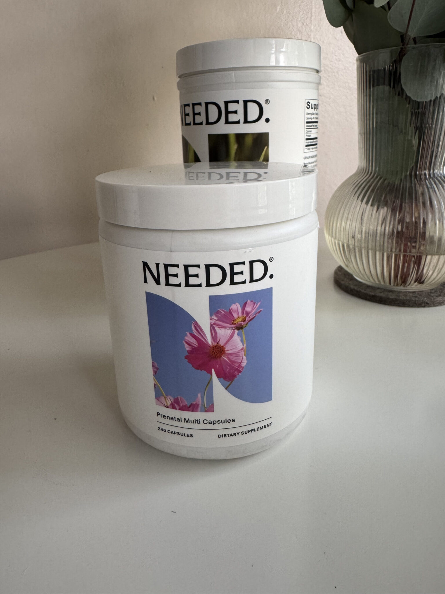 A white container labeled "NEEDED. Prenatal Multi Capsules" with a floral design on the front, placed on a white surface next to a vase and another similar container in the background.