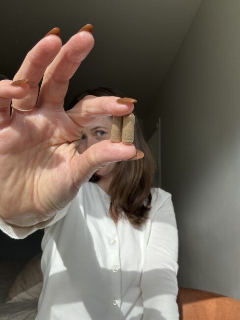 A person in a white shirt holds two brown capsules between their fingers, with their face partially obscured by their hand.